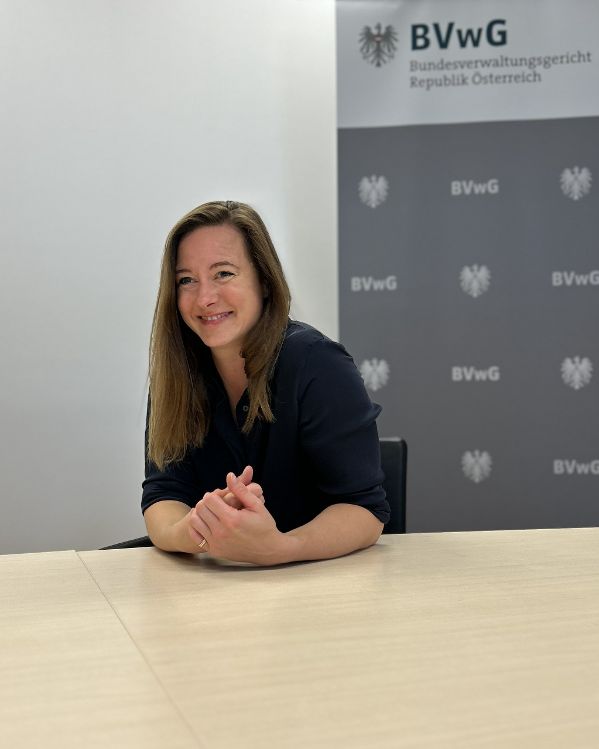 IFG-Ansprechrichterin Mag.a Julia Ludwig sitzt an einem Holztisch und spricht. Hinter ihr ist eine weiße Wand und ein Roll-Up des Bundesverwaltungsgerichts. Foto in Hochformat.