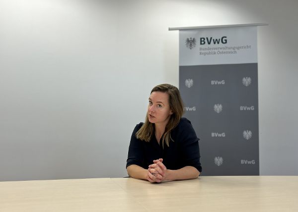 IFG-Ansprechrichterin Mag.a Julia Ludwig sitzt an einem Holztisch und spricht. Hinter ihr ist eine weiße Wand und ein Roll-Up des Bundesverwaltungsgerichts. Foto in Querformat.